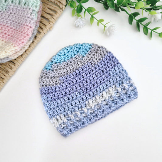 A crocheted baby beanie hat in pastel stripes of blue, purple, gray, and white, displayed on a white surface with decorative greenery and a woven mat.