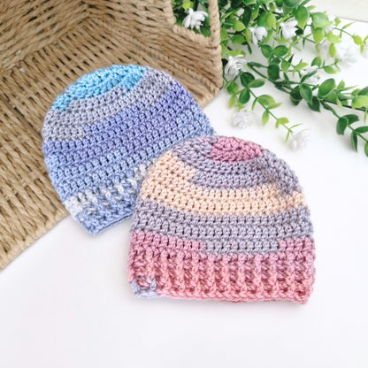 Two crocheted knit beanies displayed on a white surface with a woven basket and greenery in the background. One beanie is blue and lavender, and the other is pink, peach, and gray.
