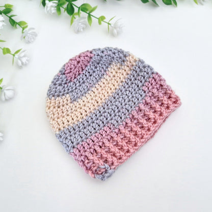 A handmade crocheted beanie hat with horizontal stripes in dusty pink, light blue-gray, and cream, styled on a white surface with decorative green leaves and small white flowers.