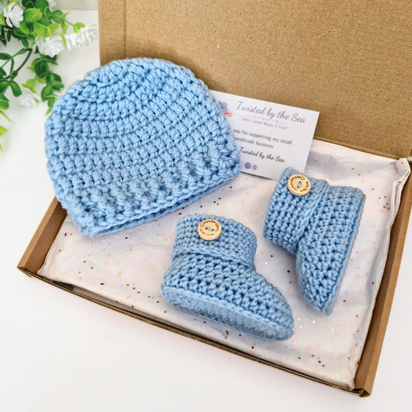 Blue crochet baby hat and booties set in a box with a card on a white background
