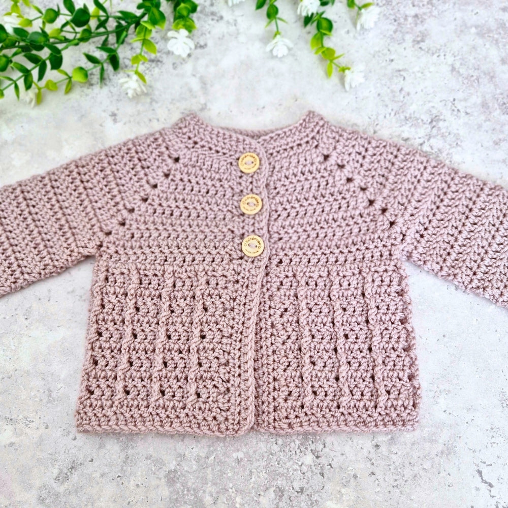 Neutral coloured crochet baby cardigan with wooden buttons on a light gray surface with greenery.