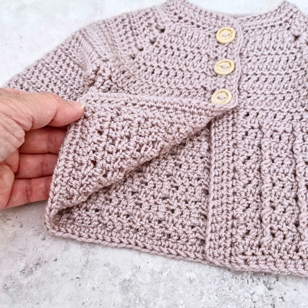 Mushroom crochet baby cardigan with button details held by a hand on a light gray surface.