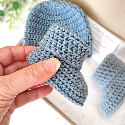 Hand holding a blue crochet baby bootie with another bootie in the background.