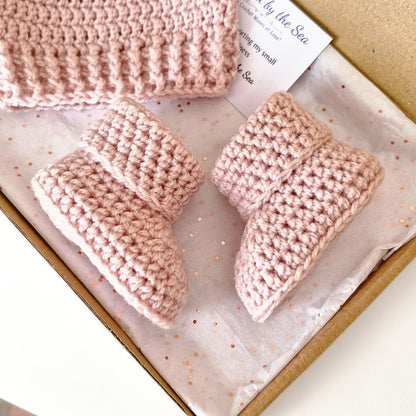 A handmade crochet baby bootie set in a gift box, including two pink crocheted booties and a matching pink knit hat, on white tissue paper with rose gold confetti.