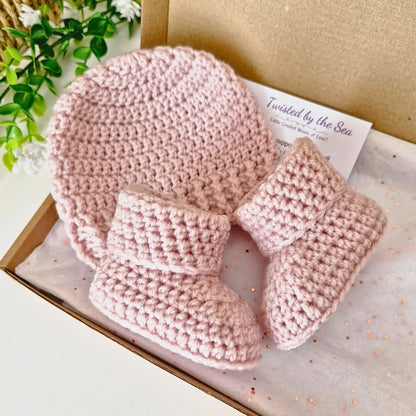 Pink crocheted baby hat and booties set in a box with a plant in the background