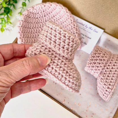 Hand holding a pink knitted baby bootie with another bootie in the background on a white surface.