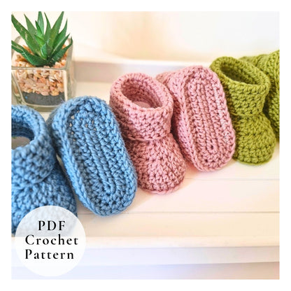Three crocheted baby booties in blue, pink, and green on a white surface with a plant in the background.