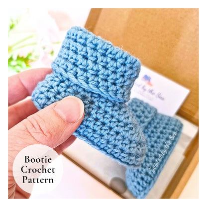 Blue crochet booties held by a hand with a pattern leaflet in the background.