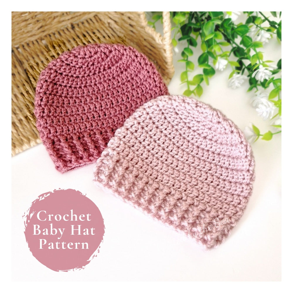 Two crocheted baby hats shown on a white background with decorative props: one dusty rose pink and one pale blush pink. A wicker basket edge and green leaves appear in the background.