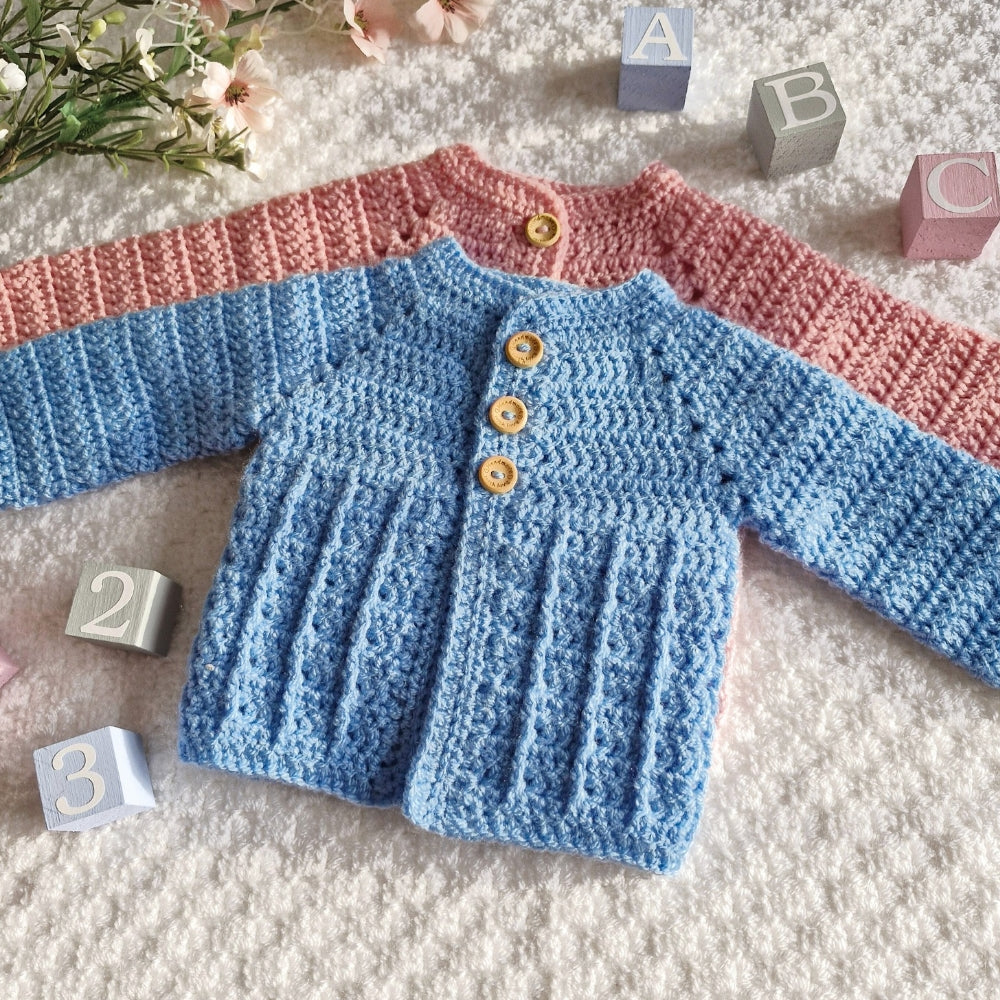 Hand-crocheted baby sweaters in pink and blue on a textured surface with blocks and flowers.