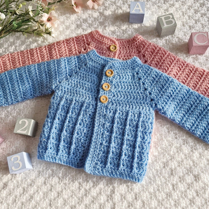 Hand-crocheted baby sweaters in pink and blue on a textured surface with blocks and flowers.