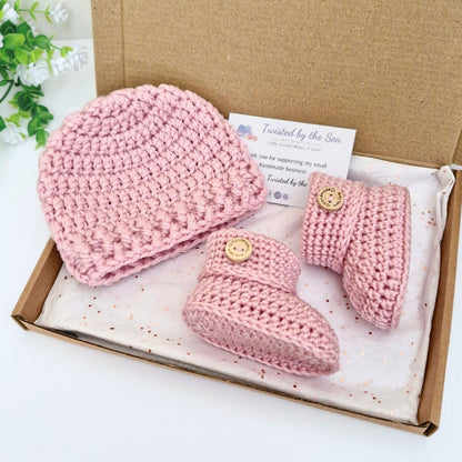 Pink crochet newborn baby hat and booties gift set in a box with a card on a white background