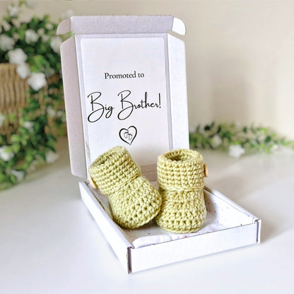 Newborn green crochet booties in a white box with 'Promoted to Big Brother!' text, on a light background.