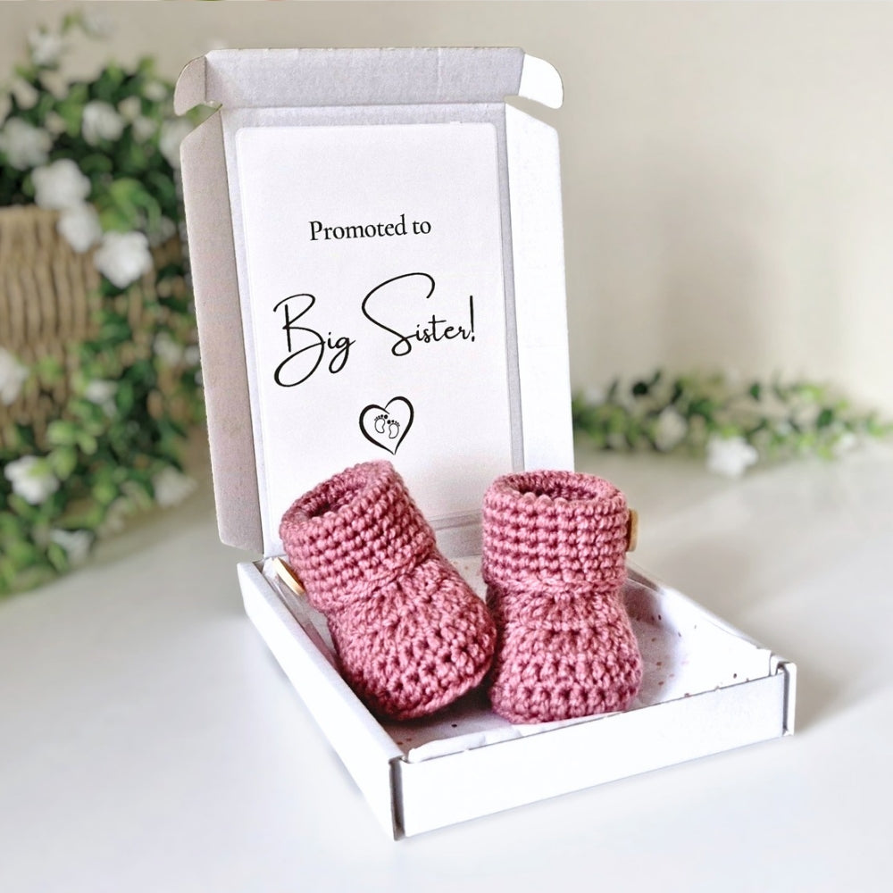 Pink crochet baby booties in a box with 'Promoted to Big Sister!' text on a light background.