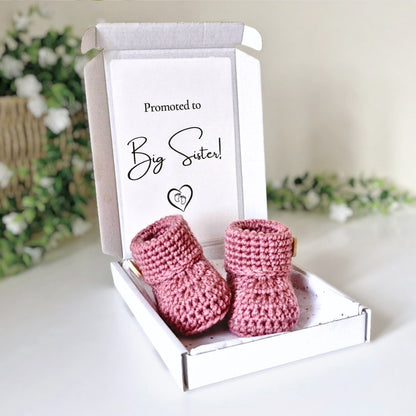 Pink crochet baby booties in a box with 'Promoted to Big Sister!' text on a light background.