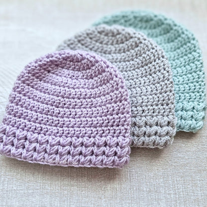 Three crocheted hats in purple, gray, and blue on a textured surface.
