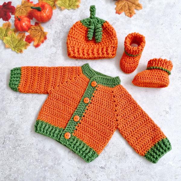 A crocheted baby outfit set styled flat on a light gray surface with autumn leaves and small pumpkins. The set includes an orange and green baby cardigan with buttons, a matching pumpkin-shaped hat, and two booties.