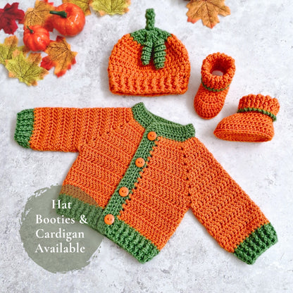 A crocheted baby outfit set arranged on a light textured surface with autumn leaves and small pumpkins. The set includes an orange cardigan with green trim and buttons, a matching pumpkin-shaped hat, and two booties.