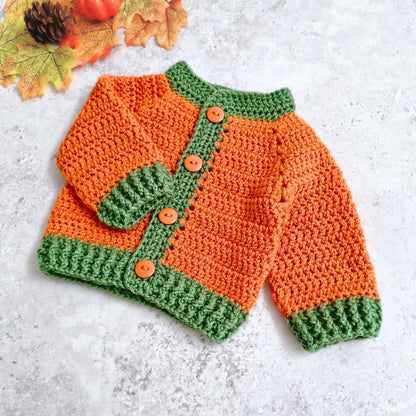 A crocheted baby cardigan with a bright orange body and green trim, featuring four orange buttons, laid flat on a light gray textured surface with autumn leaves in the corner.