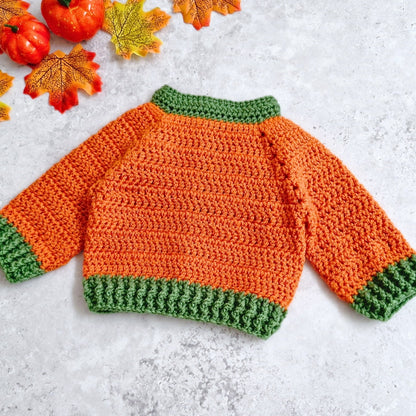 The back view of a crocheted baby sweater shaped like a pumpkin, featuring an orange body with green ribbed collar, cuffs, and hem, laid flat on a light gray textured surface with autumn leaves and small pumpkins in the corner.