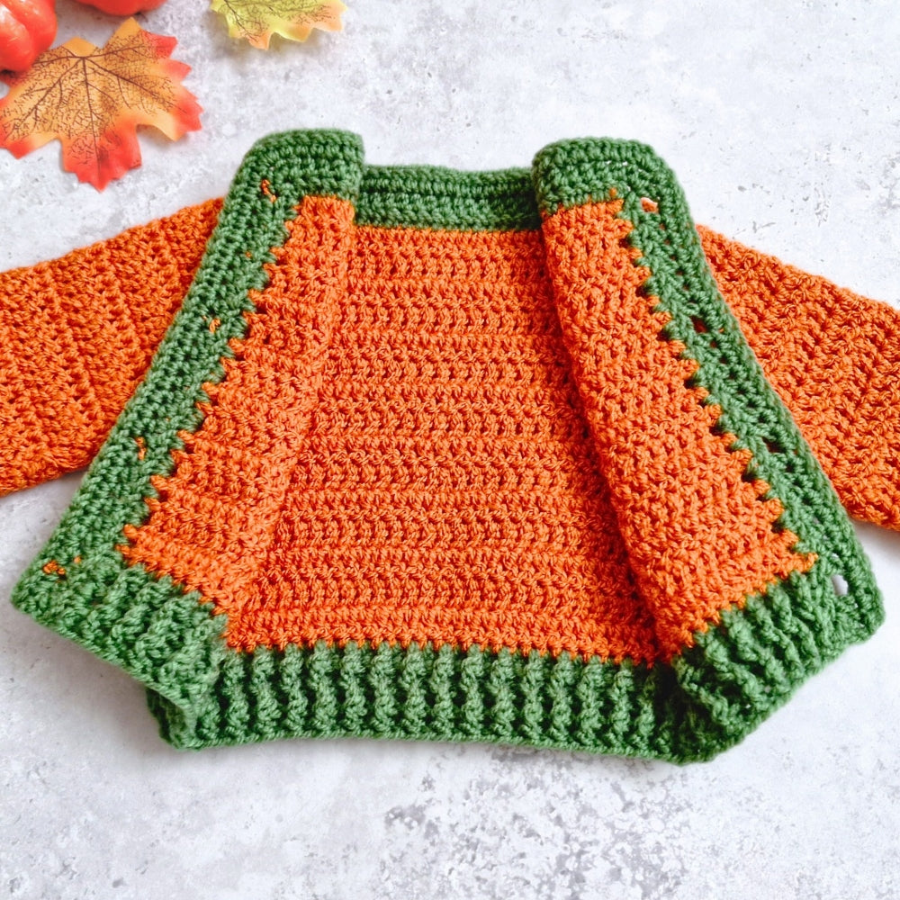 A handmade crocheted cardigan in bright orange body and green trim, laid flat on a light textured surface with autumn leaves in the corner.