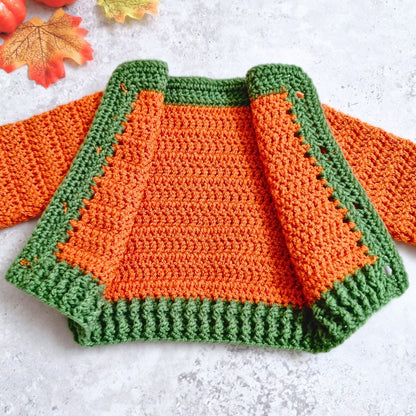 A handmade crocheted cardigan in bright orange body and green trim, laid flat on a light textured surface with autumn leaves in the corner.