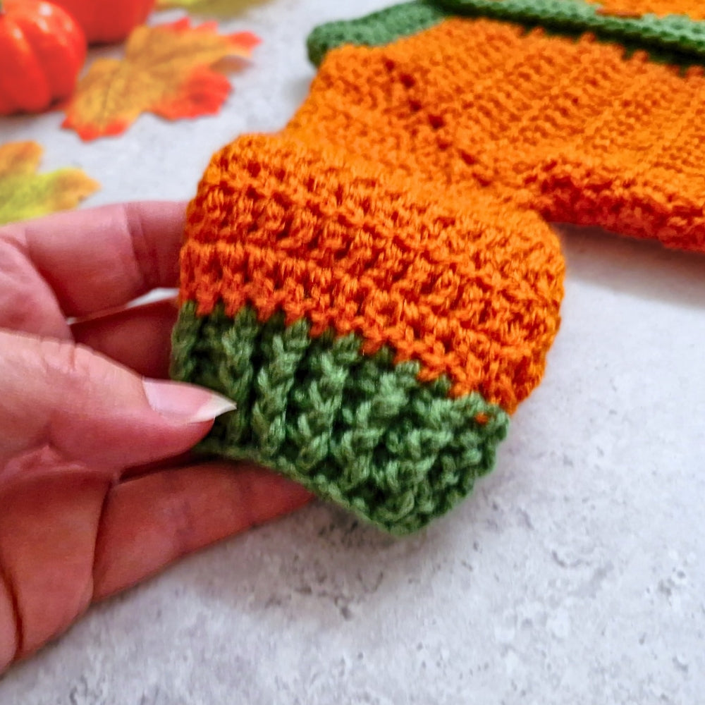 A close-up of a hand holding the sleeve of a crocheted pumpkin cardigan, in orange and green yarn. The background is a light gray surface with autumn-themed props, including orange leaves and a small pumpkin.