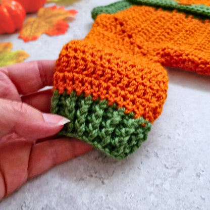 A close-up of a hand holding the sleeve of a crocheted pumpkin cardigan, in orange and green yarn. The background is a light gray surface with autumn-themed props, including orange leaves and a small pumpkin.