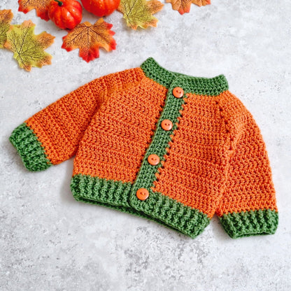 A crocheted baby cardigan styled like a pumpkin, featuring an orange body, green trim, and four orange buttons, laid flat on a light gray textured surface with autumn leaves and pumpkins in the corner. Available in sizes newborn to 2 years.