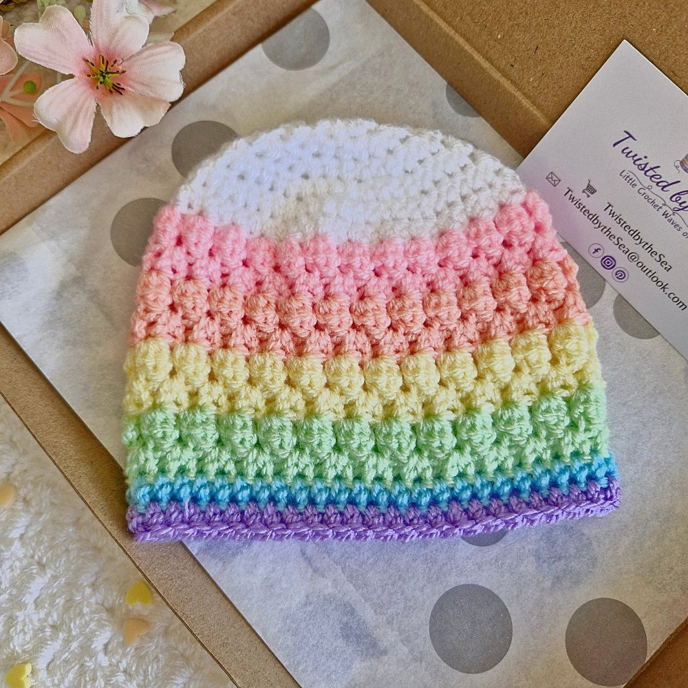 Rainbow crocheted beanie on a textured surface with a brand card in the background.