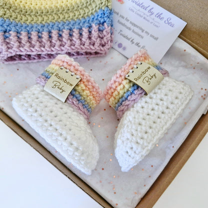 A handmade baby gift set in a cardboard box, featuring a white crocheted bootie with a pastel rainbow striped cuff.