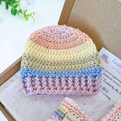 A handmade crocheted beanie hat with horizontal pastel stripes in pink, yellow, beige, blue, and lavender, displayed inside a brown gift box next to a business card.