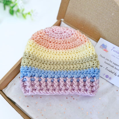 A crocheted knit beanie hat with horizontal pastel stripes in peach, yellow, beige, light blue, and lavender, displayed inside an open kraft gift box with a small card beside it.