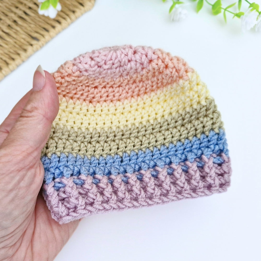 A hand holds a small crocheted beanie hat with horizontal pastel stripes in pink, peach, cream, beige, light blue, and dusty lavender on a white background.