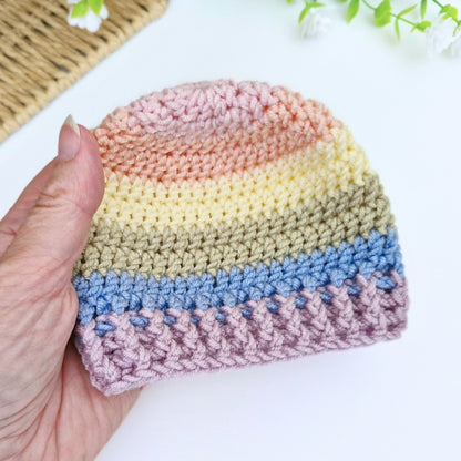 A hand holds a small crocheted beanie hat with horizontal pastel stripes in pink, peach, cream, beige, light blue, and dusty lavender on a white background.