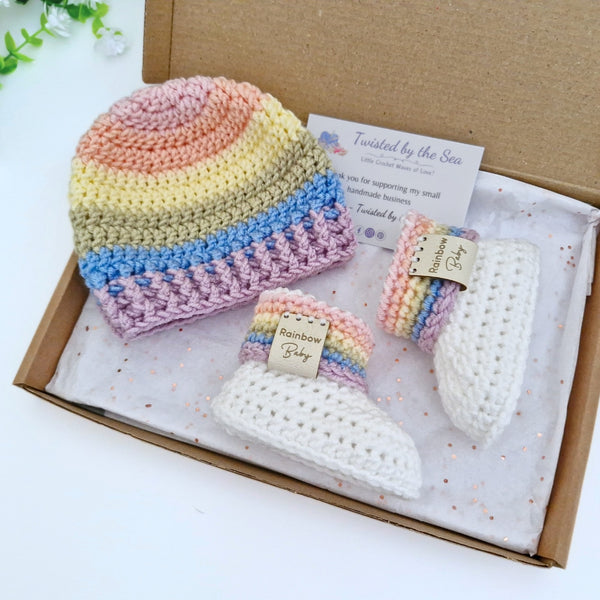 A handmade crochet baby gift set in a brown cardboard box, featuring a pastel rainbow striped beanie, a matching booties.
