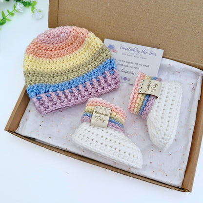 A handmade crochet baby gift set in a brown cardboard box, featuring a pastel rainbow striped beanie, a matching booties.
