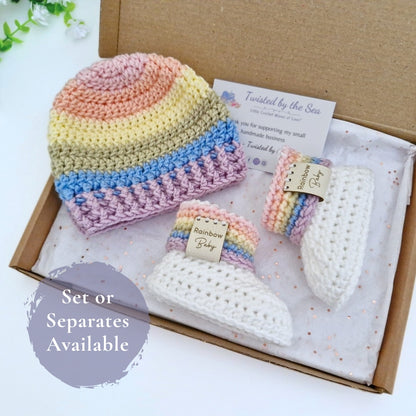 A handmade crochet baby gift set displayed inside a brown cardboard box, featuring a striped crocheted hat, and a pair of white and rainbow booties. The set includes a card from 'Twisted by the Sea' and a purple overlay that reads 'Set or Separates Available'.