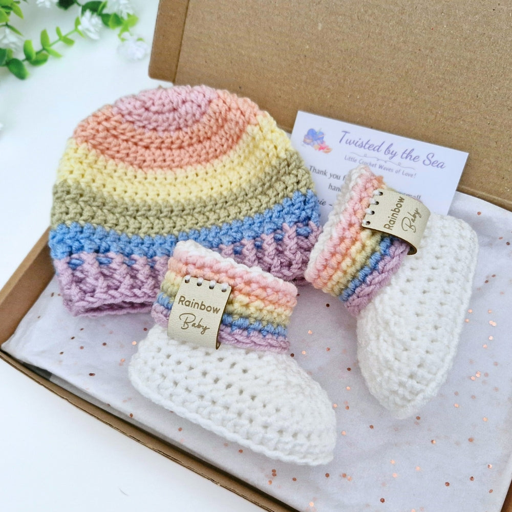 A crocheted baby gift set including a striped rainbow crochet hat, matching booties, presented in a cardboard gift box with a branded card from Twisted by the Sea.