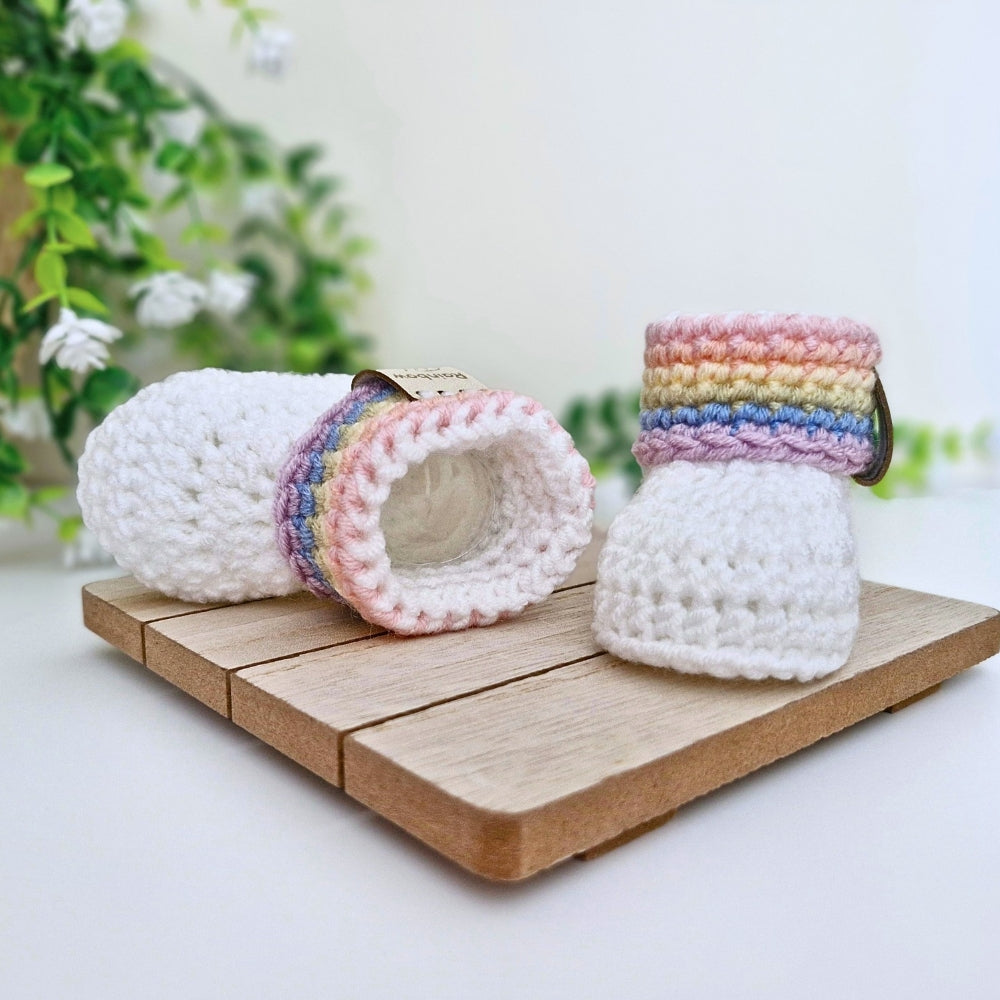 Two white crocheted baby booties with pastel rainbow striped cuffs, displayed on a small wooden block with a soft-focus floral background.
