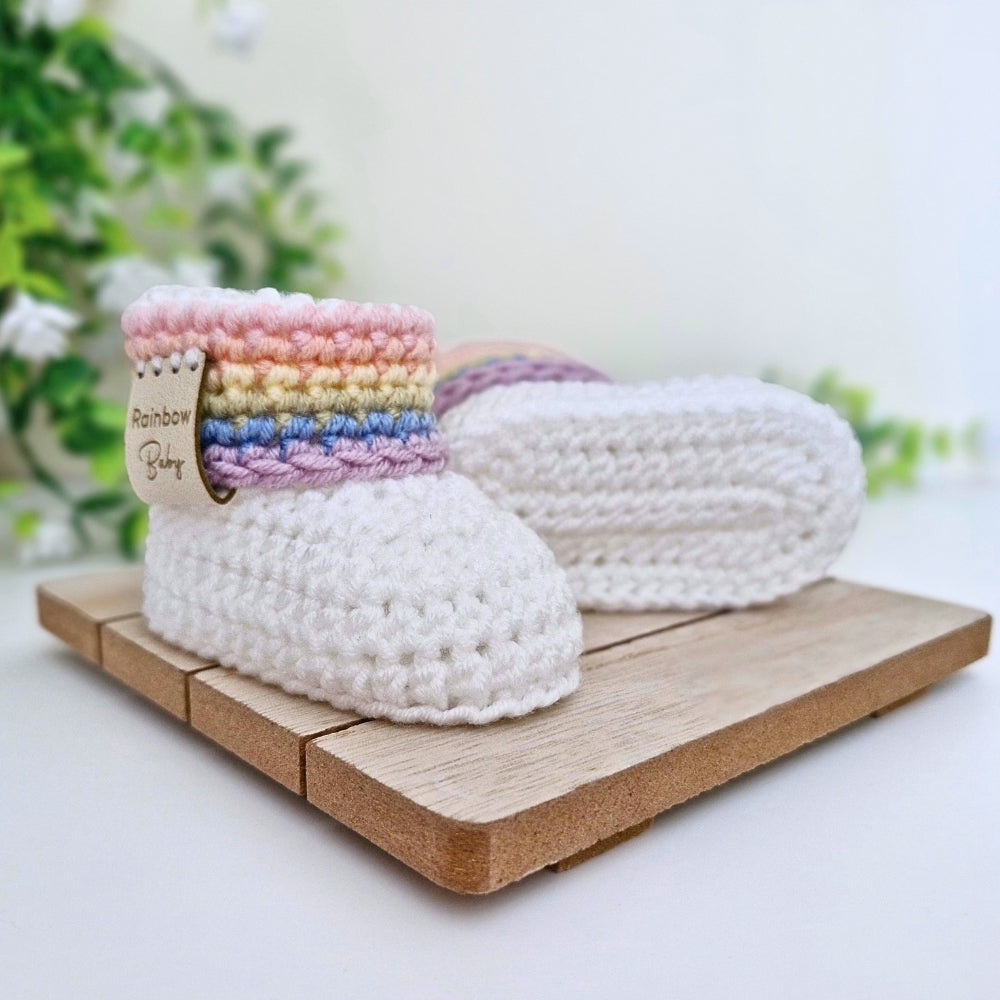 A pair of white crocheted baby booties with a pastel rainbow stripe detail on the collar, displayed on a small wooden block with a soft-focus background of green leaves.
