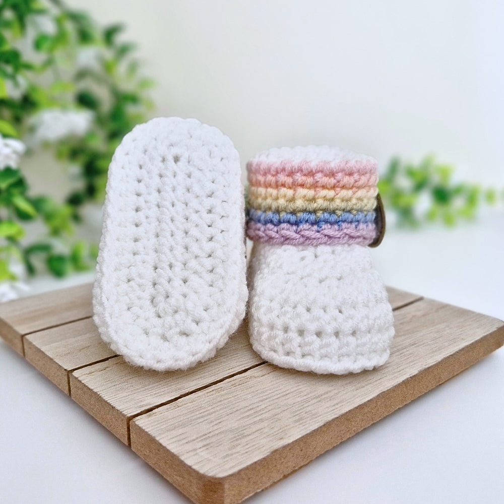 A pair of handmade crocheted baby booties, one showing the white textured sole and the other showing the top with pastel rainbow stripes, displayed on a wooden block with a soft-focus green plant background.