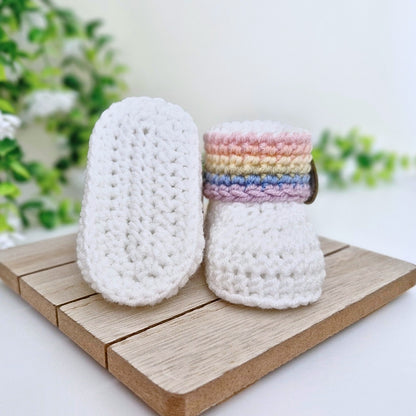 A pair of handmade crocheted baby booties, one showing the white textured sole and the other showing the top with pastel rainbow stripes, displayed on a wooden block with a soft-focus green plant background.
