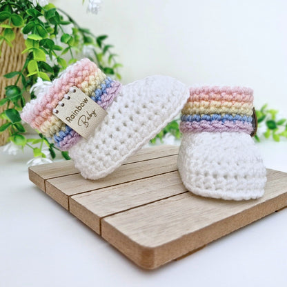 A pair of white crocheted baby booties with pastel rainbow striped cuffs, displayed on a wooden block with green plants in the background.