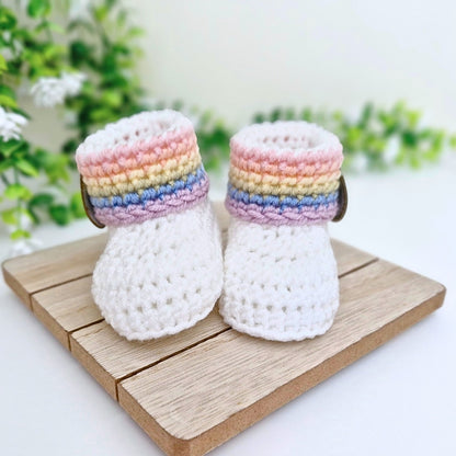 A pair of white crocheted baby booties with pastel rainbow stripes around the cuffs, displayed on a small wooden block with a soft-focus green plant background.