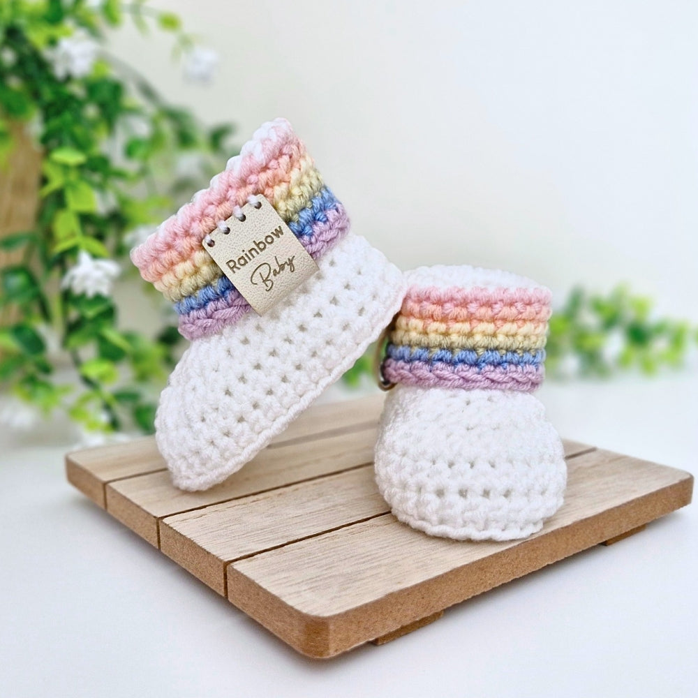 A pair of handmade crocheted baby booties in white with pastel rainbow stripes around the cuffs, displayed on a wooden block with a green plant in the background. A faux leather tag on the cuff reads 'Rainbow Baby'.