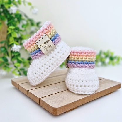 A pair of handmade crocheted baby booties in white with pastel rainbow stripes around the cuffs, displayed on a wooden block with a green plant in the background. A faux leather tag on the cuff reads 'Rainbow Baby'.