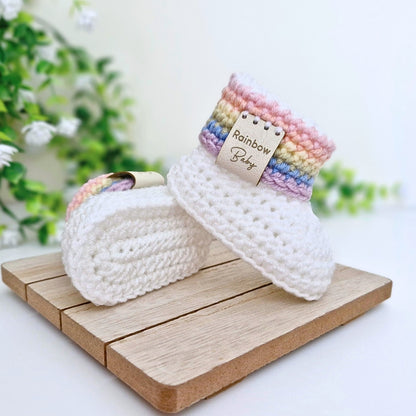 A pair of handmade crocheted baby booties in white, displayed on a small wooden block. One bootie is standing upright, showing a colorful striped cuff in shades of pink, yellow, green, blue, and purple with a small paper tag that reads 'Rainbow Baby'.