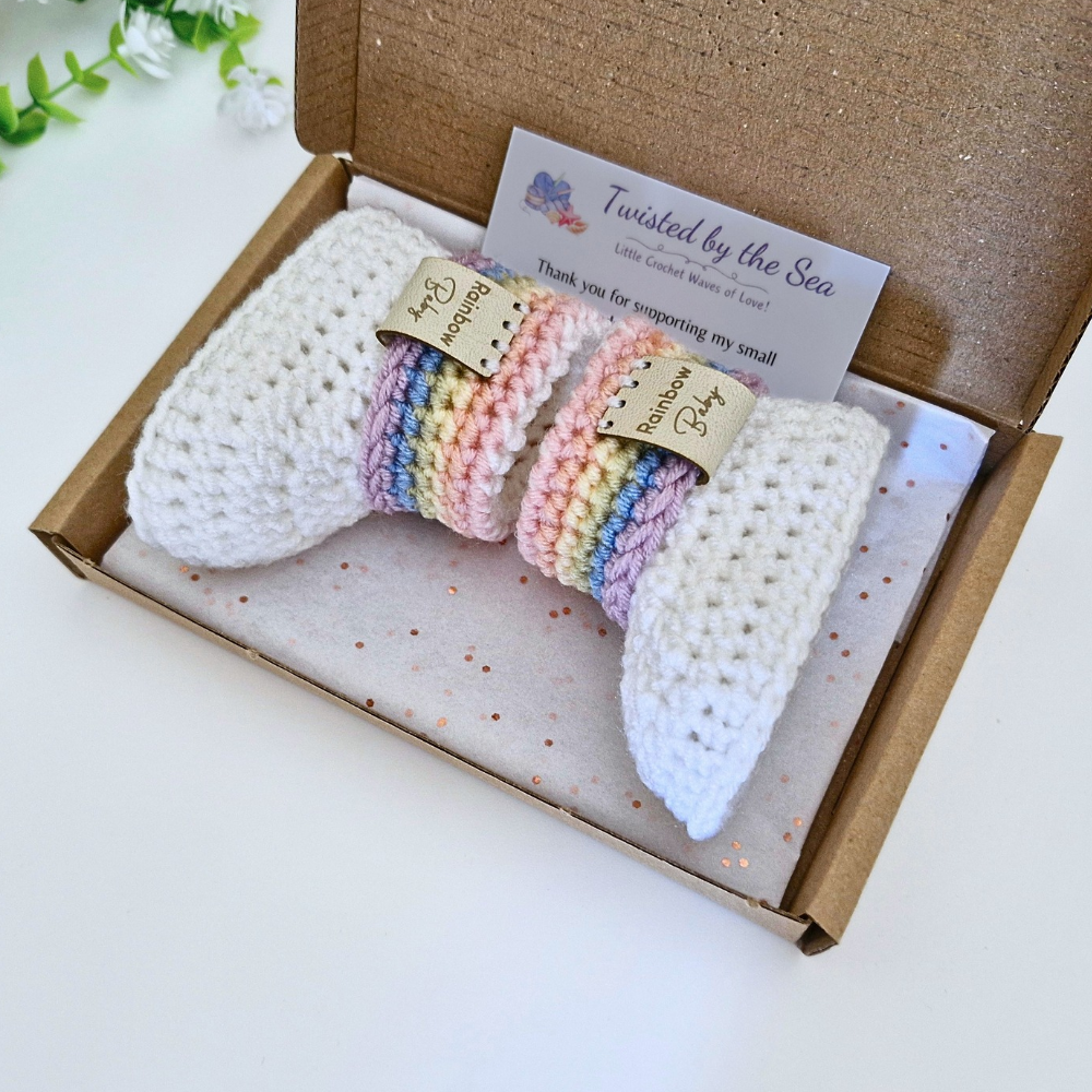 A gift box containing two white crocheted baby booties with pastel rainbow striped cuffs, displayed on a white surface with a glittery brown box and a thank-you card from Twisted by the Sea.