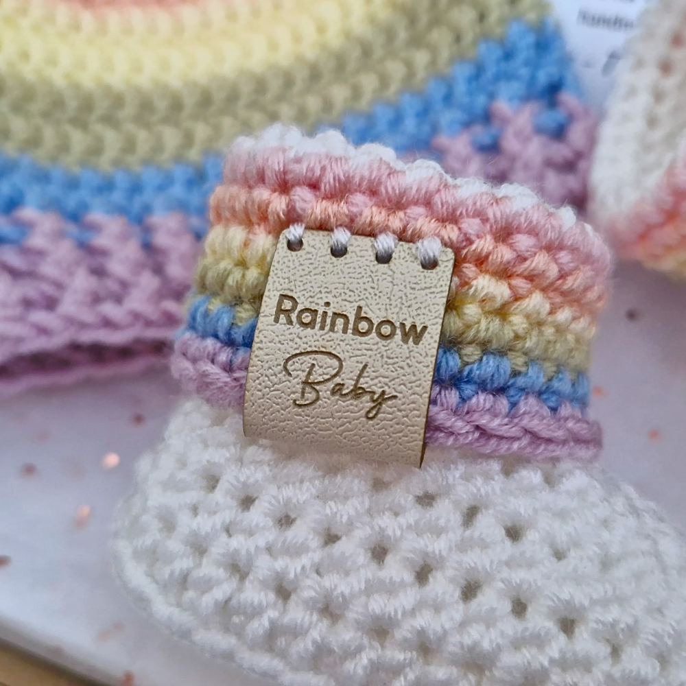 A close-up of a handmade crocheted baby bootie with pastel rainbow stripes in pink, yellow, blue, and purple, featuring a beige faux-leather strap with a 'Rainbow Baby' faux leather tag.